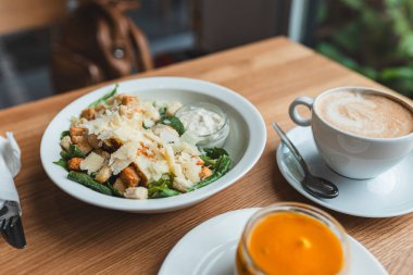 İçinde kızarmış ekmek, tavuk ve parmesan peyniri olan beyaz bir kase dolusu Cezar salatası. Cezar salatası ve bir fincan kahve beyaz tabakta, ahşap masanın üzerinde. Öğle yemeği için farklı yemekler.