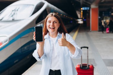 Kırmızı bavullu gülümseyen beyaz kadın tren istasyonunda elinde boş ekranlı akıllı telefon tutuyor. Online Wi-Fi internet konsepti, istasyon platformunda bekleyen mutlu hipster