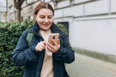 Sırt çantalı neşeli bir kadın şehir sokaklarında iletişim kurmak için akıllı telefon aleti tutuyor. Açık havada dinlenirken cep telefonu tutan kadın, şehir yaşam tarzı konsepti.