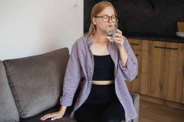 Evdeki kanepede oturup su içen neşeli bir kadın. Sağlıklı yaşam alışkanlığı, yaşam tarzı konsepti