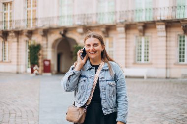 Şehir caddesinden akıllı telefonlu mutlu genç bir kadın arıyor. Teknoloji, iletişim konsepti. 30 'lu yaşlardaki pozitif kadın şehir geçmişinde telefonla konuşuyor.