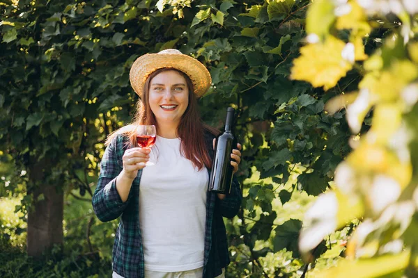Woman tasting red wine in vineyard. Portrait of pretty young woman holding glass of wine and bottle of wine. Mockup for design