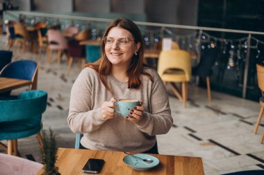 Gözlüklü çekici bir kadın, tatlı bir gülümseme içinde kahve içerken arada dinleniyor. Rahat giyinen, gülümseyen ve rahat bir kafede bir bardak sıcak içecekle dinlenen pozitif bir kadın. Kahvaltı kavramı.