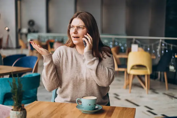 Angry casual woman calling on the phone in a cafe. Female feels upset and mad while having conversation on her phone. Person sitting at table and using smartphone