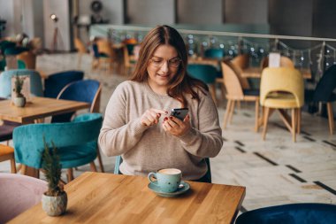 Gülümseyen ve kafede akıllı telefon kullanan genç ve mutlu bir kadın. Masada oturan ve kapalı alanda akıllı telefon kullanan biri. Çevrimiçi eğitim, düzen, alışveriş kavramı