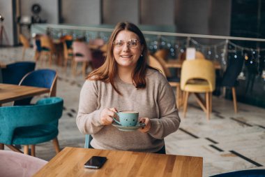 Gözlüklü çekici bir kadın, tatlı bir gülümseme içinde kahve içerken arada dinleniyor. Rahat giyinen, gülümseyen ve rahat bir kafede bir bardak sıcak içecekle dinlenen pozitif bir kadın. Kahvaltı kavramı.
