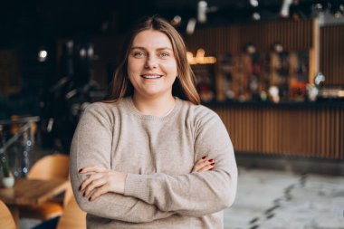 Kollarını kavuşturup kameraya bakan genç bir kadının portresi. Kafede duran mutlu kız