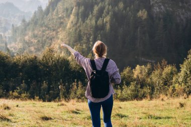 30 'lu yaşlardaki sarışın kız, dağların arka planını işaret ediyor. Sırt çantalı genç bir kadın doğa yürüyüşü yapıyor. Spor kıyafetlerle yürüyen güzel bir kadın. Etkin yaşam tarzı kavramı