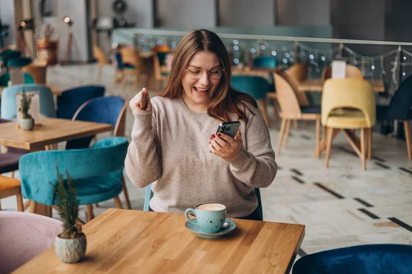 Satisfied young woman holding phone and celebrating in a cafe. hipster female in glasses overjoyed with winning online contest on web site