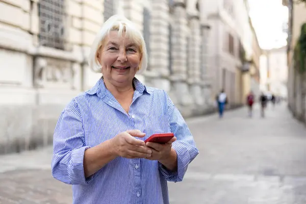 Happy senior with phone walking on city street. Mature blonde woman using smartphone on city street. Urban lifestyle concept.