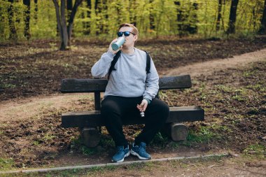 Spor elbiseli mutlu sarışın adam yürüyüş yaptıktan sonra duruyor. 30 'ların Yürüyüşçüsü su şişesinden su içer ya da mavi termostan sıcak içecek