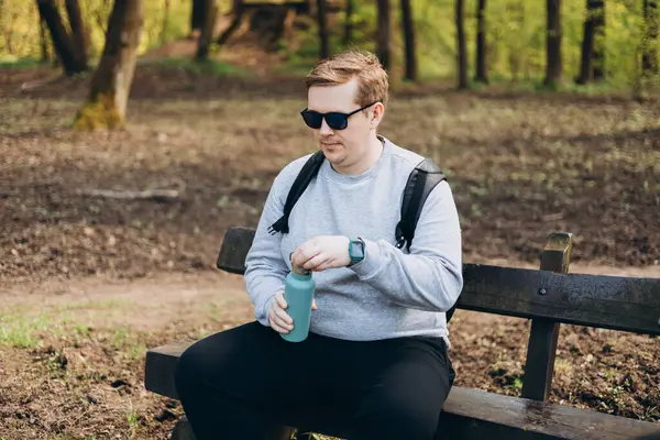 Happy blonde man in sport clothes having a halt after hiking. 30s Hiker drinking water from water bottle or hot drink from blue thermos