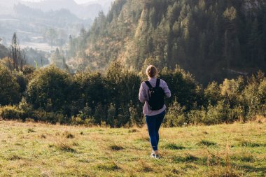 Otuzlu yaşların kızı dağ zirvesinde yeşil çimlerle kaplı sırt çantasıyla yazın gün batımında sisli dağ vadisine bakıyor. Sportif genç bayanla manzara