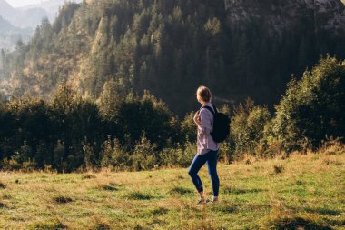 Otuzlu yaşların kızı dağ zirvesinde yeşil çimlerle kaplı sırt çantasıyla yazın gün batımında sisli dağ vadisine bakıyor. Sportif genç bayanla manzara