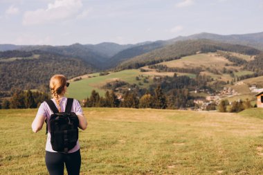 Otuzlu yaşların kızı dağ zirvesinde yeşil çimlerle kaplı sırt çantasıyla yazın gün batımında sisli dağ vadisine bakıyor. Sportif genç bayanla manzara