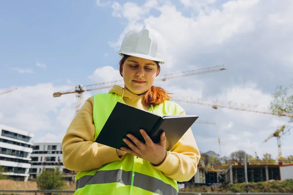 Happy Architect with documents at a construction site. Portrait of successful woman constructor wearing white helmet and safety yellow vest. Woman are planning building project.