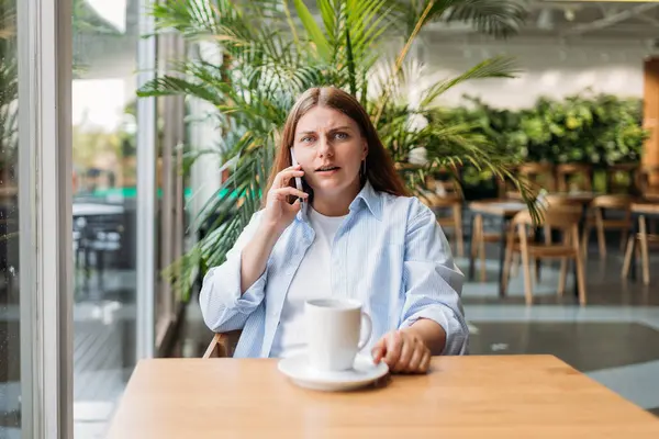 Upset woman talking over phone disappointed hear bad news, sad pensive girl speaking having unpleasant phone conversation and sitting in a cafe at a table by the window