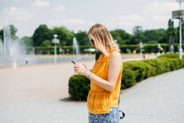 Sırt çantalı neşeli sarışın kadın şehir caddesinde yürürken akıllı telefonunu kontrol ediyor. Açık havada akıllı telefon kullanan 30 'lu yaşlardaki güzel bir kızın portresi. Kentsel yaşam tarzı konsepti.