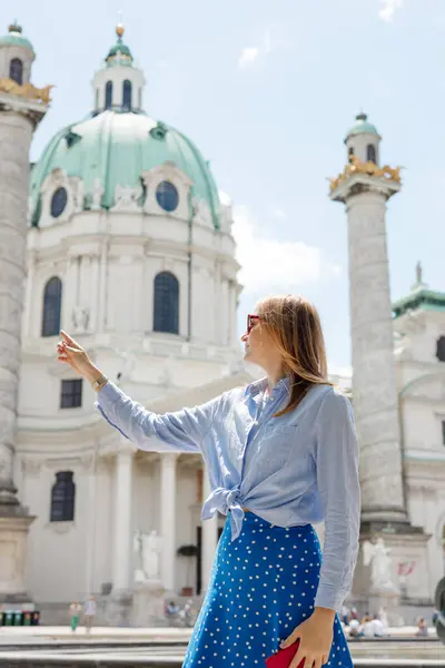 Young female tourist in sunglasses pointing finger near Catholic Church Karlskirche, Vienna. Saint Karl Cathedral. Traveling Europe in summer time. High quality photo