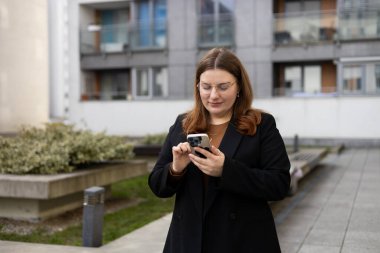 Modern şehir caddesinde güneşli bir günde gülümseyen bir kadın akıllı telefondan mesaj atıyor. Başarılı küçük işletmeler.