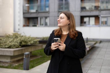 Modern şehir caddesinde güneşli bir günde gülümseyen bir kadın akıllı telefondan mesaj atıyor. Başarılı küçük işletmeler.