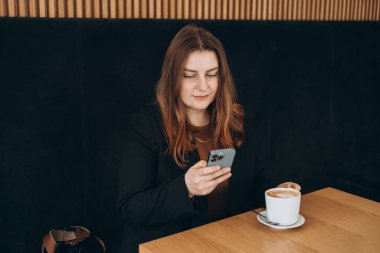 Genç mutlu iş kadını bir kafede gülümsüyor ve akıllı telefon kullanıyor. Masada oturan ve kapalı alanda akıllı telefon kullanan biri. Çevrimiçi eğitim, düzen, çalışma kavramı