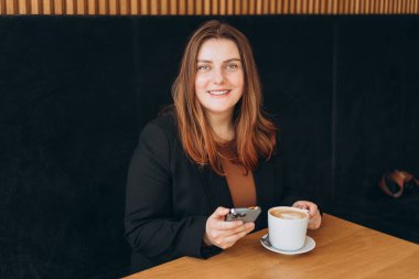 Genç mutlu iş kadını bir kafede gülümsüyor ve akıllı telefon kullanıyor. Masada oturan ve kapalı alanda akıllı telefon kullanan biri. Çevrimiçi eğitim, düzen, çalışma kavramı