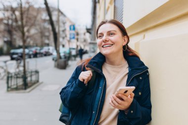 Sırt çantalı mutlu genç kadın akıllı telefonunu kontrol ediyor. Açık havada akıllı telefon kullanan 30 'lu yaşlardaki güzel bir kızın portresi. Şehir ya da teknoloji kavramı.