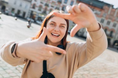 30 'lu yaşlarda kadın, şehir dışında parmaklarından yapılmış bir çerçeveye bakıyor. Şehir caddesinde yürüyen neşeli genç bir kadın, şehir yaşam tarzı konsepti.