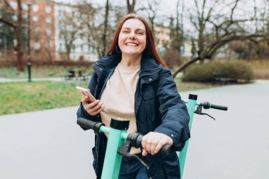 Elektrikli scooter 'ın yanında duran gülümseyen genç kadın akıllı telefon kullanıyor. Ekolojik nakliye. Etkin yaşam biçimi.