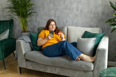 Günlük giysiler içinde neşeli genç bir kadın kanepede oturur ve evdeki oturma odasında vakit geçirir. Dinlen, keyfine bak, keyfine bak, keyfine bak. 30 'lu yaşlarda kadın. Cep telefonu kullanıyor. Kredi kartı yok.