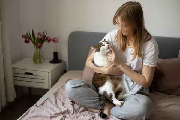 stock image Young blonde woman is resting with a cat on the bed at home one spring day. Morning at home. Cute cat licking fingers of female owner in daytime at home