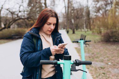Elektrikli scooter 'ın yanında duran gülümseyen genç kadın akıllı telefon kullanıyor. Ekolojik nakliye. Etkin yaşam biçimi.