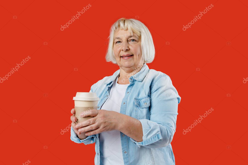 Mature blonde woman hold craft paper brown cup coffee isolated on red background, studio portrait