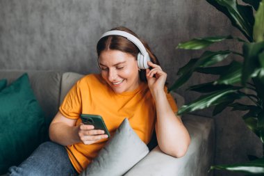 Gülümseyen kız evde dinleniyor, akıllı telefon kullanarak müzik çalıyor ve beyaz kulaklık takıyor. Genç bir kadın evdeki kanepede iyi ses sesinden zevk alıyor..