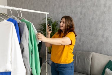 Elbiselerini satmak ya da bağışlamak için akıllı telefon kullanan bir kadın gömleğin fotoğrafını çekiyor. Deklutating, Sıralama Kıyafetler. Tekrar kullan, ikinci el konsept. Bilinçli tüketici.