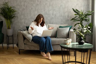 Laptop kullanan ve kötü haberler okuyan üzgün genç bir kadın, evde oturan üzgün düşünceli bir kız. Negatif insanların duyguları. Oturma odasında çalışan kızıl saçlı kadın.