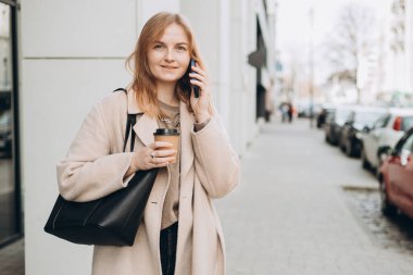 Moda kıyafetleri içindeki neşeli, genç sarışın kadın telefonda bir fincan çayla konuşuyor. Mockup, elinde akıllı telefon ve kahve fincanı tutan kadın. Şehir konsepti