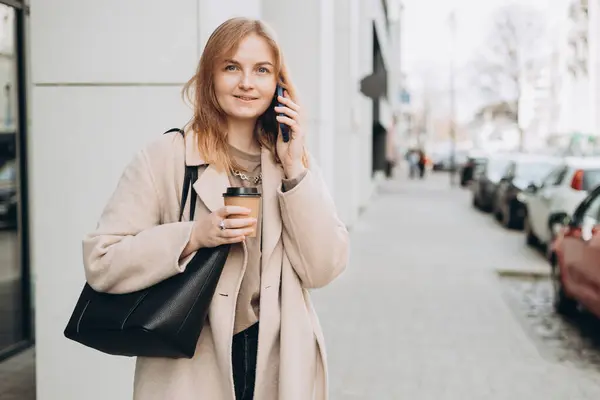 Happy cheerful young blonde woman in fashion clothes talking on Phone with eco cup of tea. Mockup, Woman holding smartphone and coffee cup. Urban concept