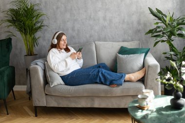 Gülümseyen kız evde dinleniyor, müzik dinliyor, akıllı telefon kullanıyor ve beyaz kulaklık takıyor. Genç bir kadın evdeki kanepede iyi ses sesinden zevk alıyor.