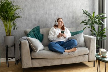 Gülümseyen kız evde dinleniyor, müzik dinliyor, akıllı telefon kullanıyor ve beyaz kulaklık takıyor. Genç bir kadın evdeki kanepede iyi ses sesinden zevk alıyor.