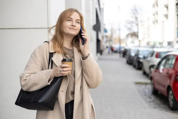 Happy cheerful young blonde woman in fashion clothes talking on Phone with eco cup of tea. Mockup, Woman holding smartphone and coffee cup. Urban concept