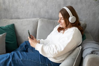 Gülümseyen kız evde dinleniyor, müzik dinliyor, akıllı telefon kullanıyor ve beyaz kulaklık takıyor. Genç bir kadın evdeki kanepede iyi ses sesinden zevk alıyor.