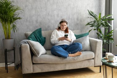 Gülümseyen kız evde dinleniyor, müzik dinliyor, akıllı telefon kullanıyor ve beyaz kulaklık takıyor. Genç bir kadın evdeki kanepede iyi ses sesinden zevk alıyor.