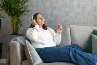 Gülümseyen kız evde dinleniyor, müzik dinliyor, akıllı telefon kullanıyor ve beyaz kulaklık takıyor. Genç bir kadın evdeki kanepede iyi ses sesinden zevk alıyor.