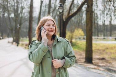 Parkta telefonla konuşan gülümseyen güzel bir kadının portresi. 30 'lu yaşlardaki neşeli kadın turist, günlük kıyafetlerini giymiş, dışarıda dinleniyor.