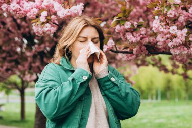 Parkta çiçek açan ağaçların arasında burun sileceği olan genç kızıl saçlı bir kadın. Hasta kadınların portresi beyaz dokuya hapşırır, burun kıvrımlarından muzdariptir. Soğuk algınlığı veya alerji belirtileri.