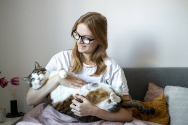 Evde kedisi olan mutlu genç bir kadın. Soğuk havada, evcil hayvan dostu konsept. Sokak kedisi yatakta uyuyor. Rahat ev arka planı, sabah yatma zamanı..