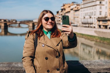 Çekici genç bayan turist yeni şehri keşfediyor. Floransa 'daki ünlü Old Bridge' de elinde akıllı telefon tutan kızıl saçlı kız. Seyahat ediyorum. Mutlu kız şehirde yürüyor ve selfie çekiyor.