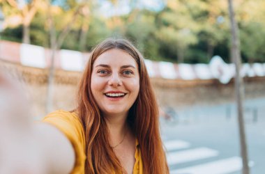 Şehirde yürürken selfie çeken gülümseyen genç bir kadın. Sosyal medya uygulamasında paylaşmak için dışarıda poz veren neşeli kız öğrenci. Beyaz kadın.